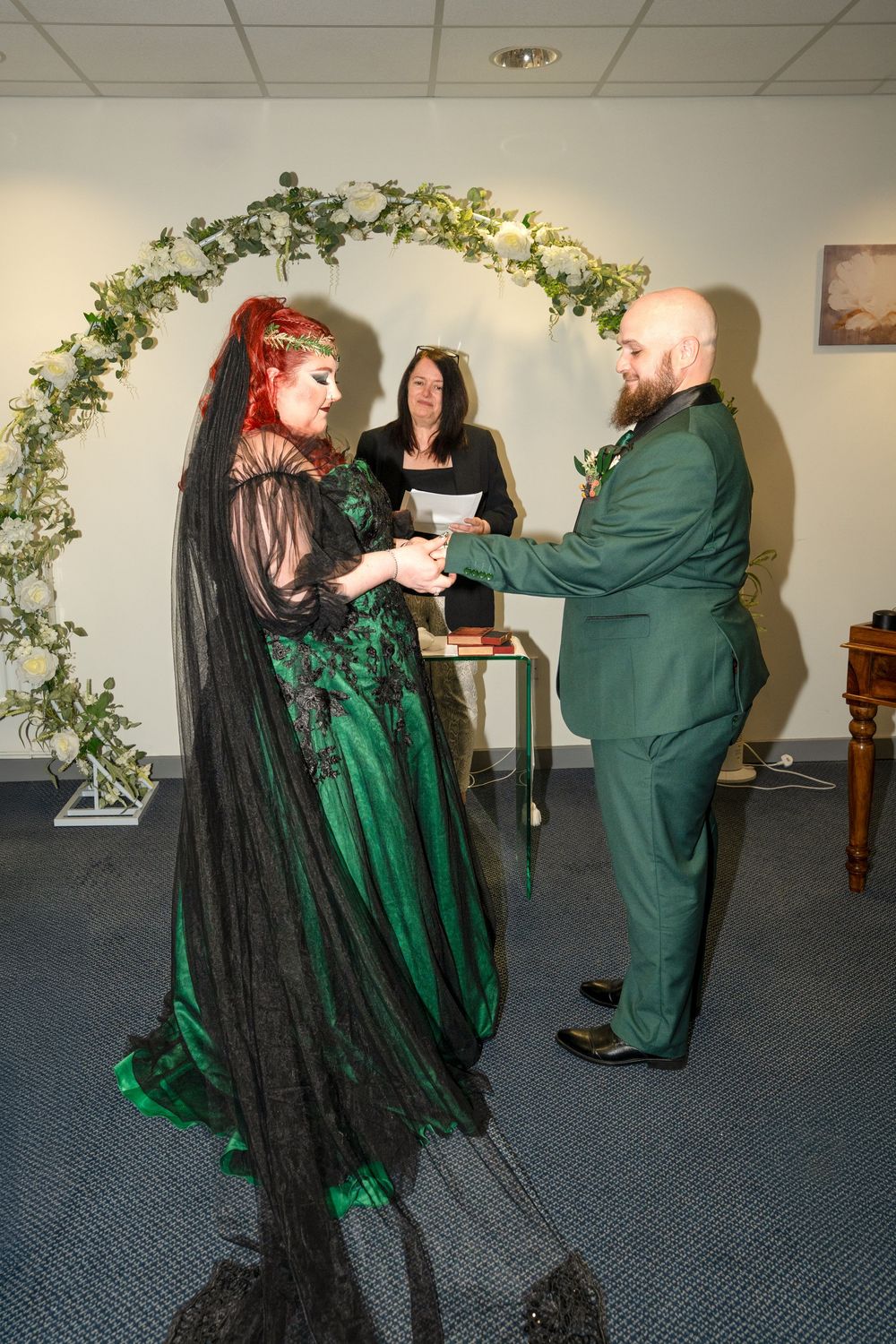 Gothic wedding couple in green and black attire exchange vows under floral arch with officiant.