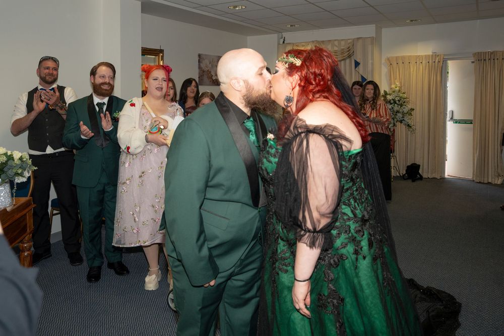 Newlywed couple shares first kiss in green wedding attire surrounded by applauding guests at indoor ceremony.