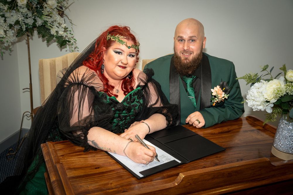 Couple in green and black wedding attire signing marriage certificate at wooden desk with floral decor.