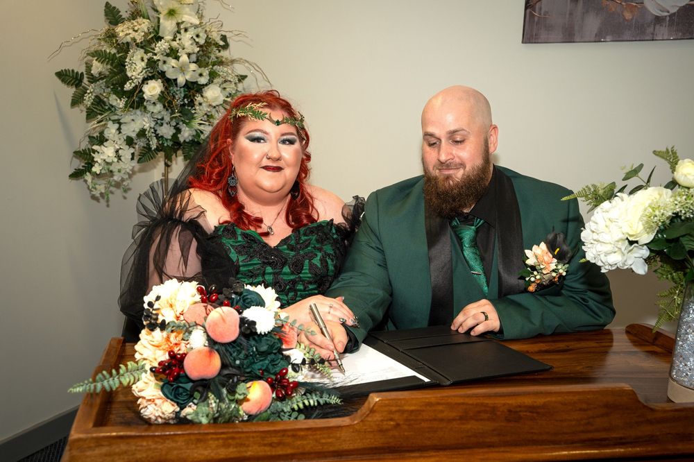 Couple in green wedding attire signing marriage documents at a decorated table with floral arrangements.