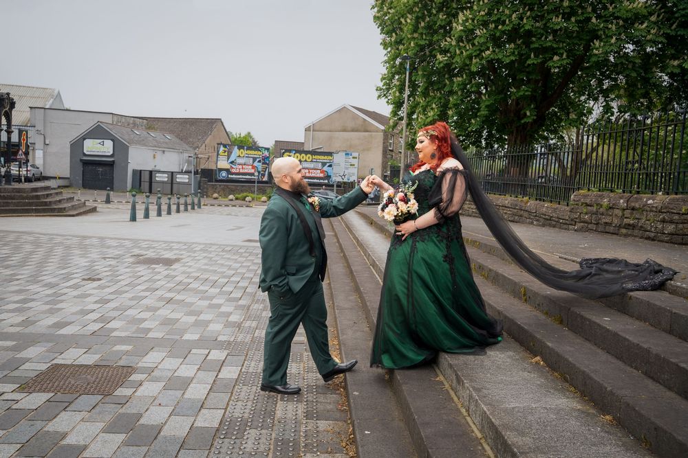 Couple in emerald green wedding attire holding hands on a stone plaza, bride with long red hair and flowing veil.