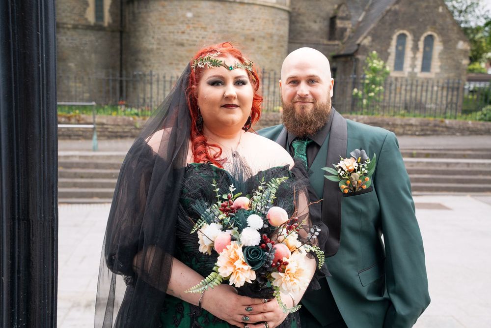 Bride in black gothic wedding dress with red hair and groom in green suit pose outside a church.