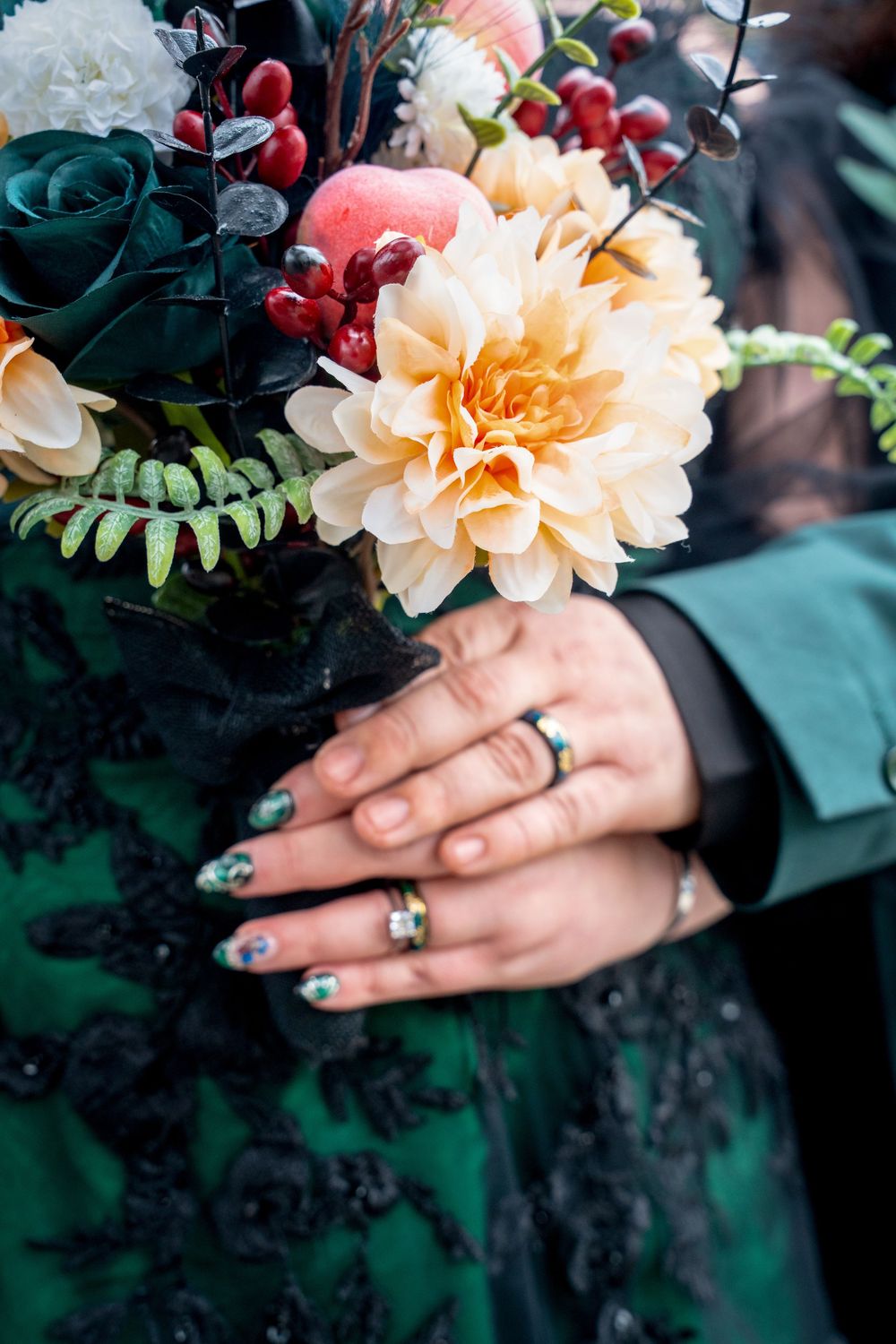 Bride and groom hands holding a bouquet with dahlias, roses, and berries, wearing matching green gemstone rings.