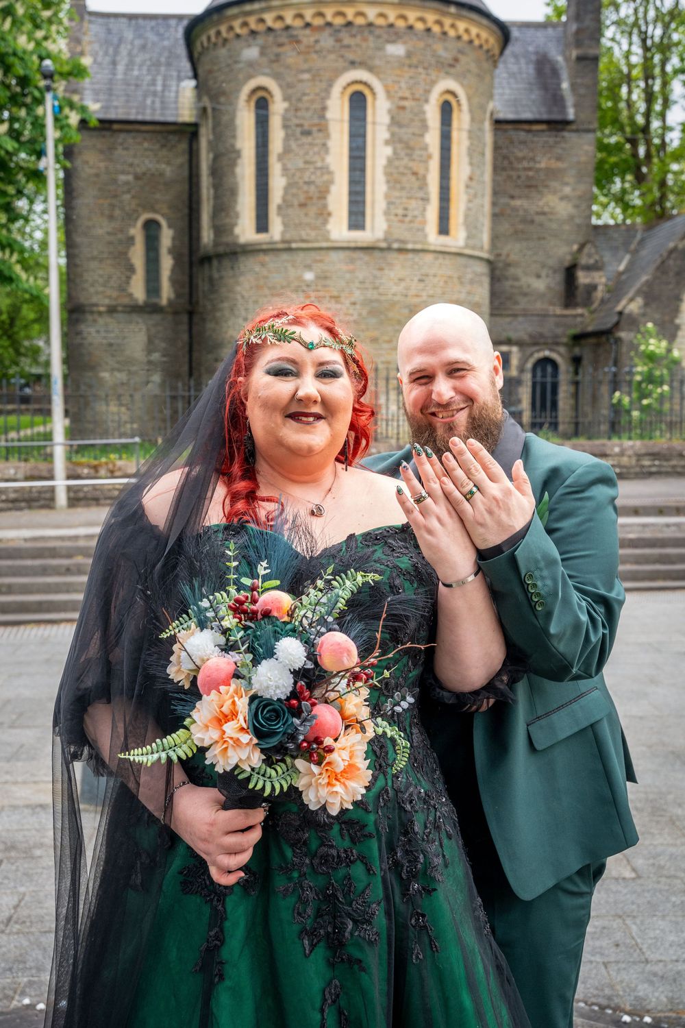 Newlywed couple in green and black wedding attire showing rings outside a stone church, bride holds bouquet.