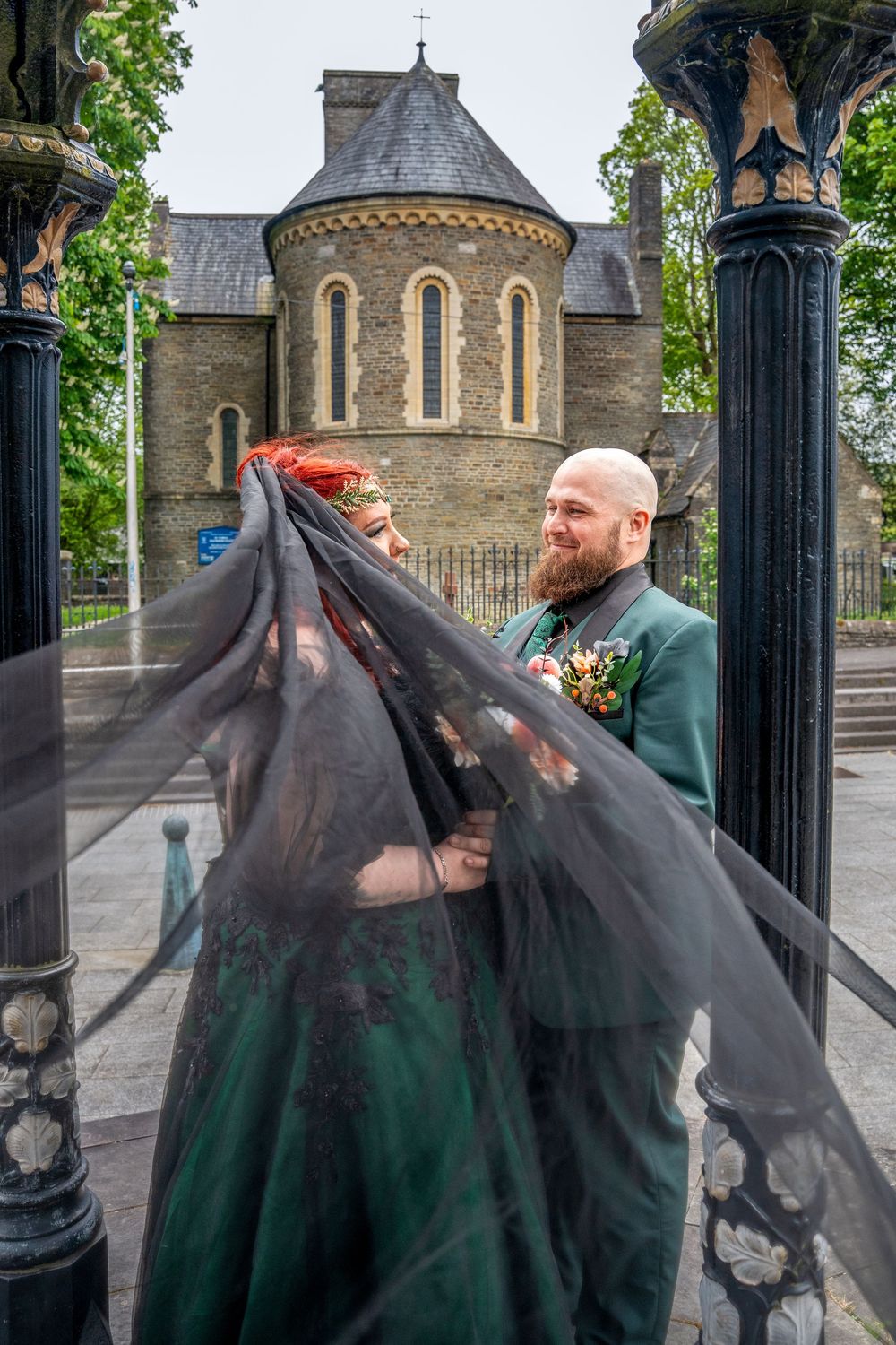 Couple in gothic wedding attire with black veil near historic stone church, groom in green suit holding bouquet.