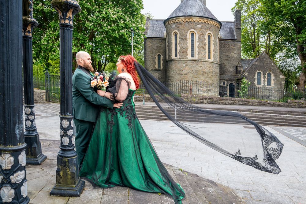 Couple in green wedding attire embracing outdoors, bride in emerald gown with black veil, groom in teal suit.