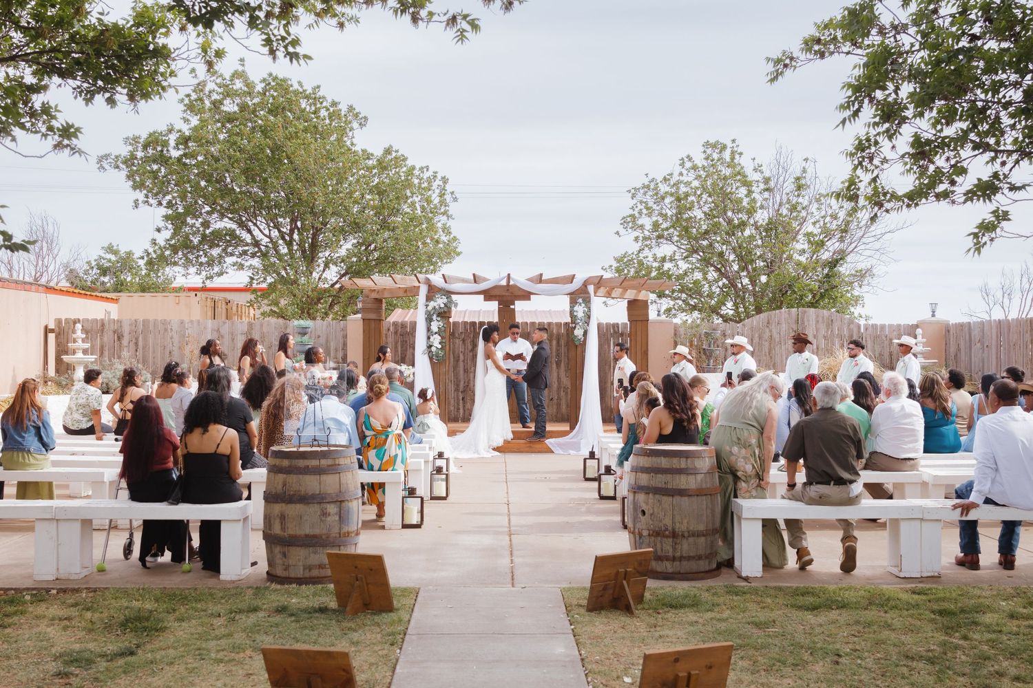 Outdoor rustic wedding ceremony with guests seated on white benches and barrels, couple at wooden arch altar.