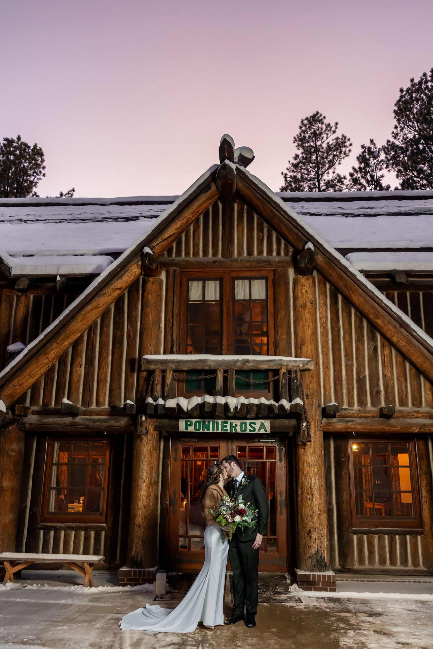 Couple kisses in front of snowy Ponderosa log cabin venue at dusk during winter wedding.