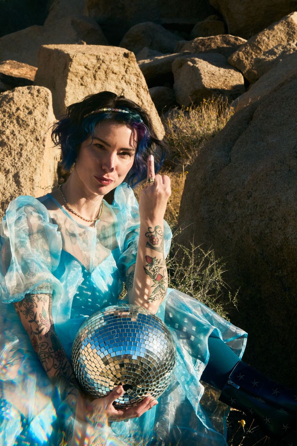 Tattooed woman in blue organza dress holding disco ball among desert rocks.
