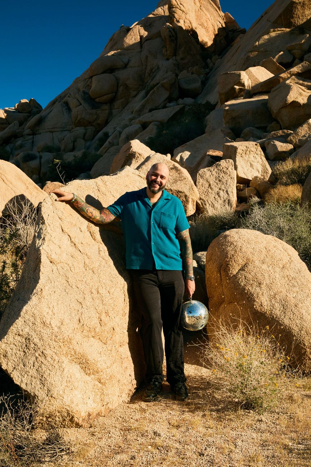 Smiling bald man with tattoos holding a disco ball beside large boulders in a desert landscape under blue sky.