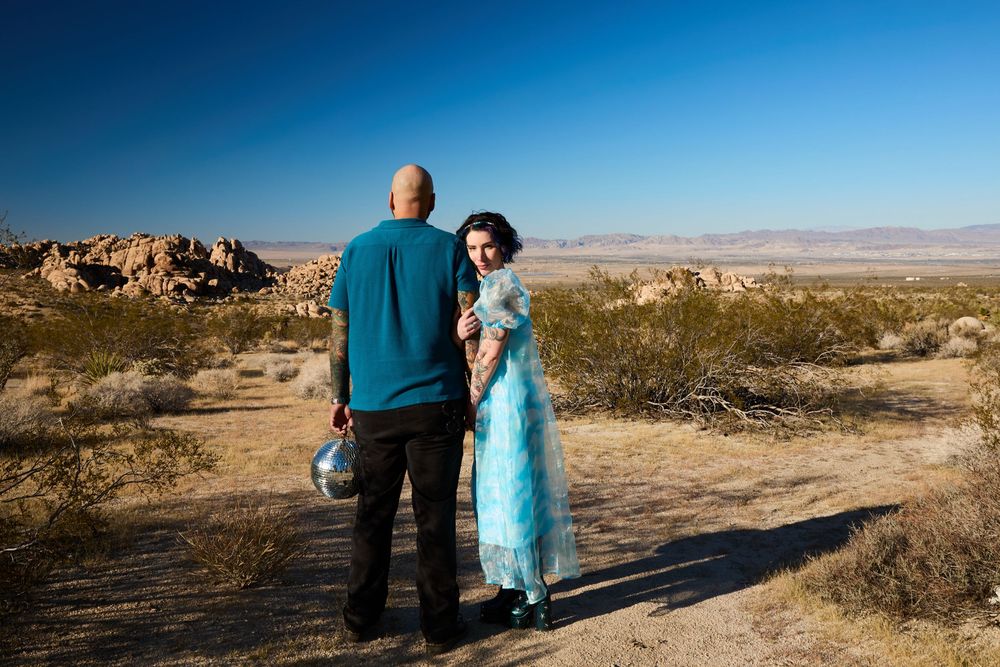 A bald man and woman in a blue gown stand on a desert path surrounded by rocky landscape under a clear blue sky.