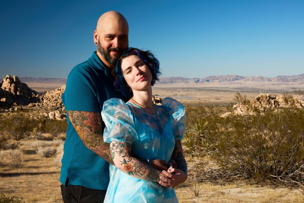 Couple embracing in desert landscape with mountains, woman in blue dress, man with tattoos, Joshua Tree backdrop.