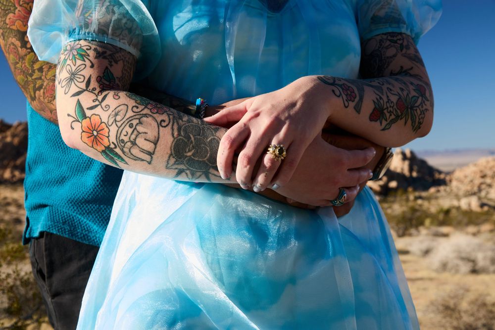 Couple embracing outdoors, showing tattooed arms and a gold ring, wearing blue outfits in a desert landscape.