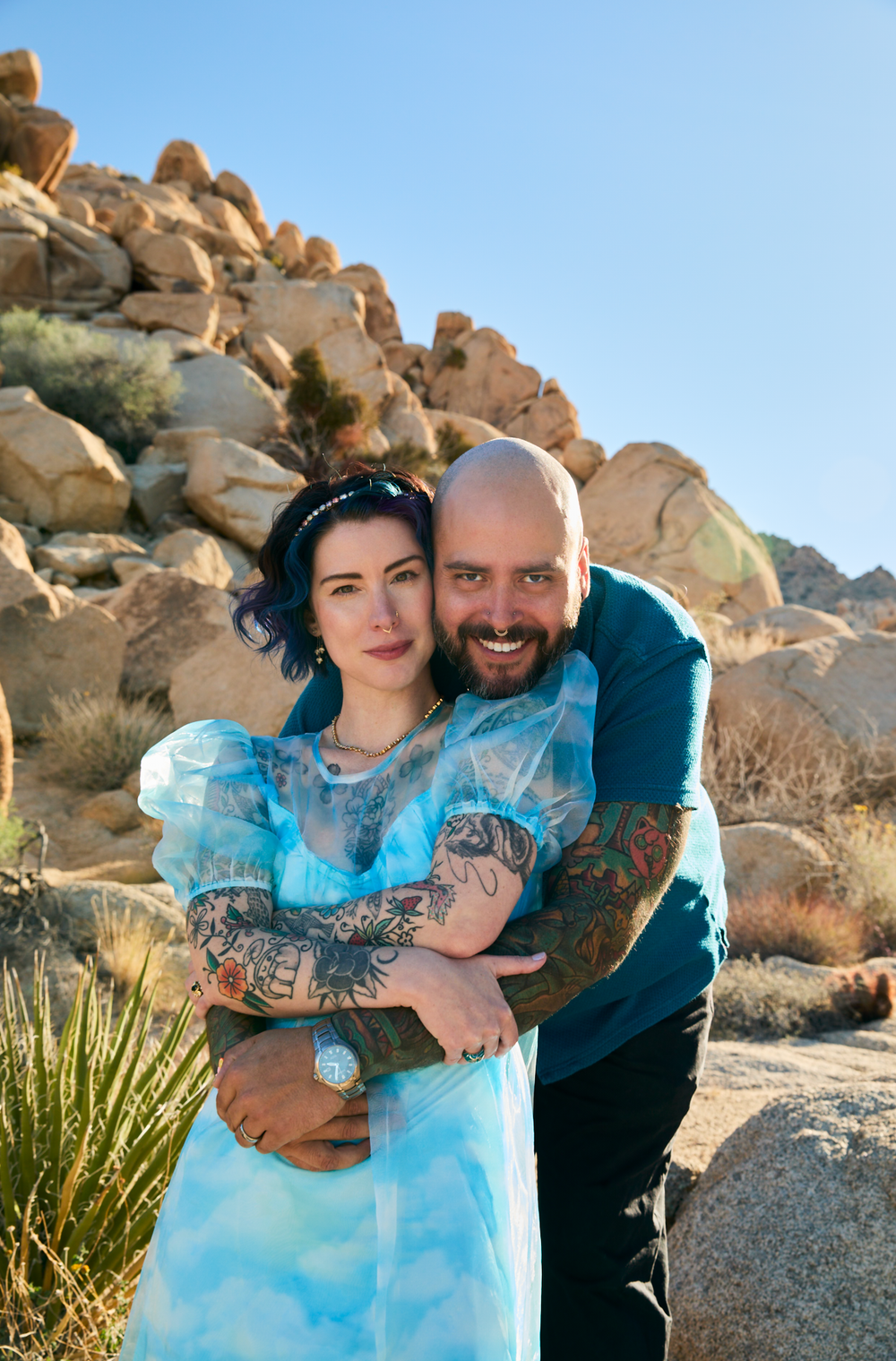 A tattooed couple embraces and smiles in Joshua Tree desert landscape, woman in blue dress with purple hair, bald bearded man.
