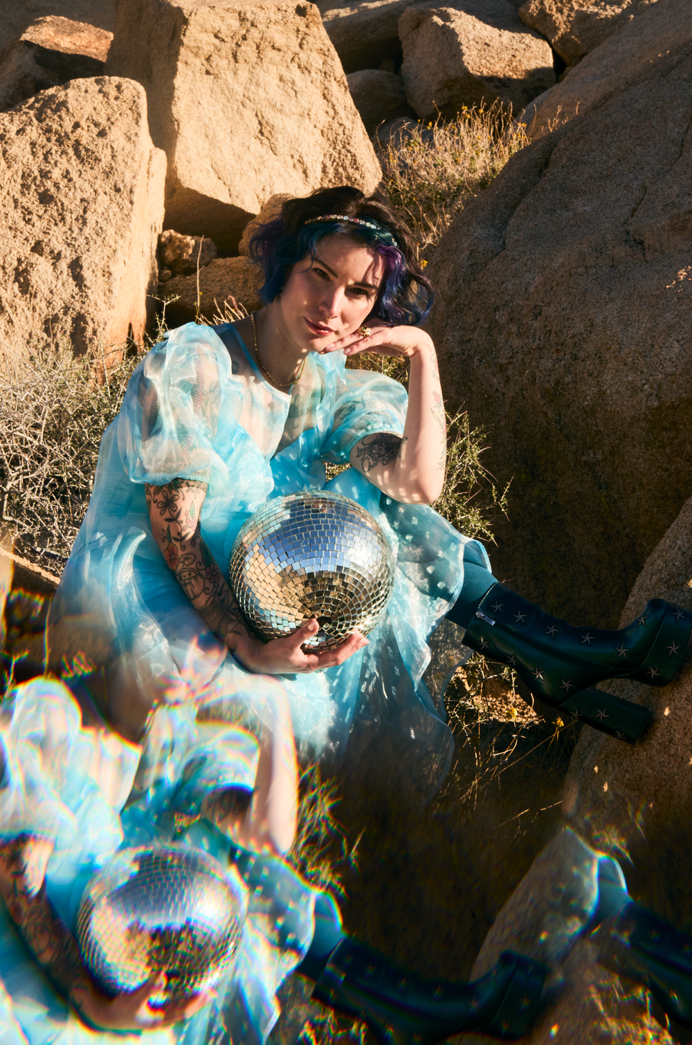 Woman in blue tulle dress holding disco ball among desert rocks, purple hair, tattoos, editorial fashion photography.