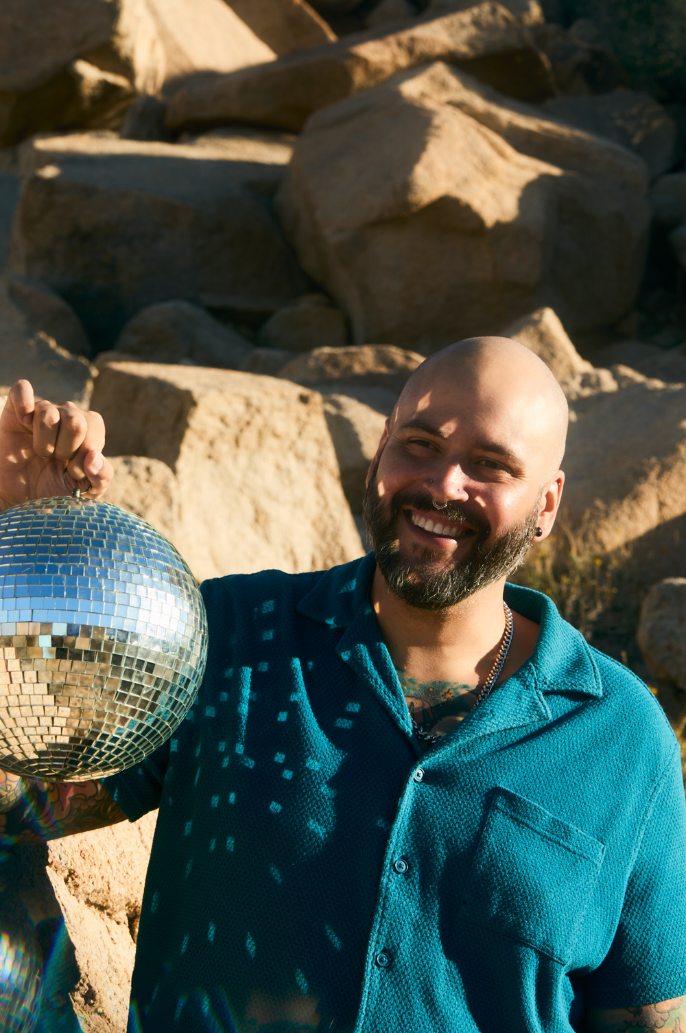 Smiling bald man with beard holding a disco ball outdoors near rocky boulders, wearing a teal polo shirt.