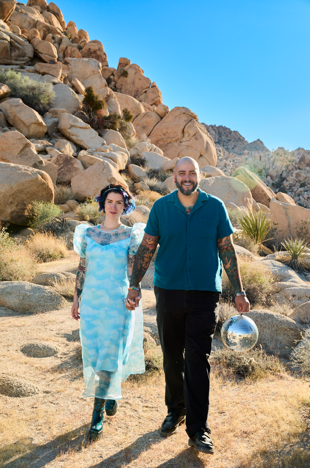 Tattooed couple walking hand-in-hand through Joshua Tree desert landscape, woman in blue dress, man in teal shirt holding disco ball.