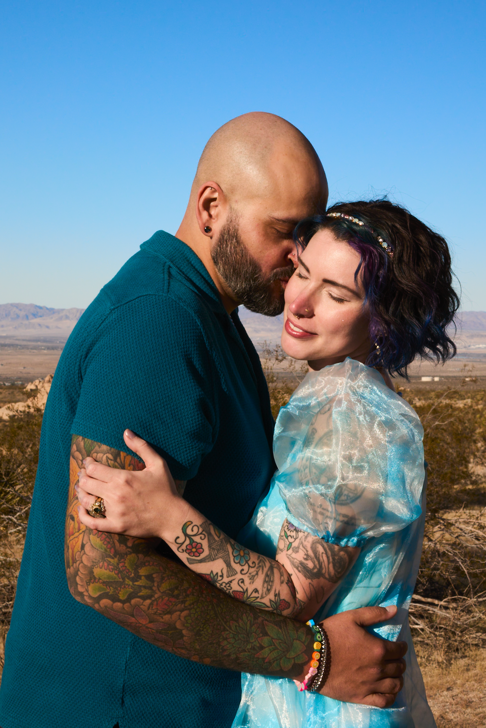 Tattooed couple embracing outdoors in desert landscape, man kissing woman's forehead, blue sky background.