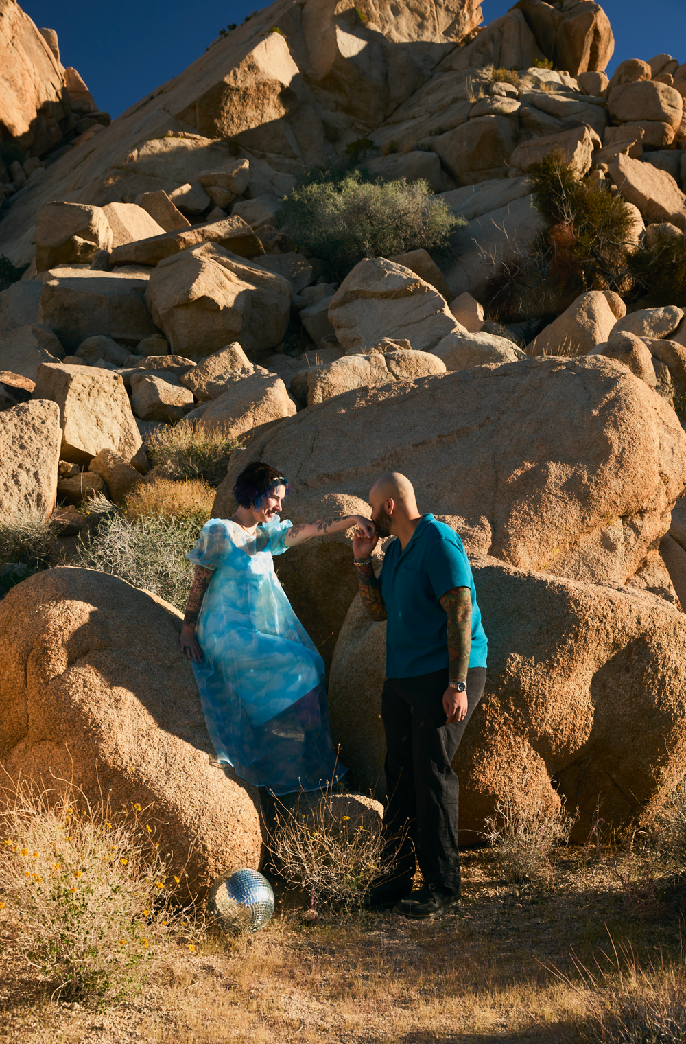 A bald man in teal and a woman in blue dress interact among large boulders in a desert landscape.