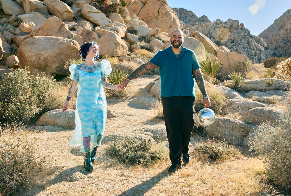 Couple holding hands walking on a desert trail among boulders, woman in blue dress, man in teal shirt holding bowl.