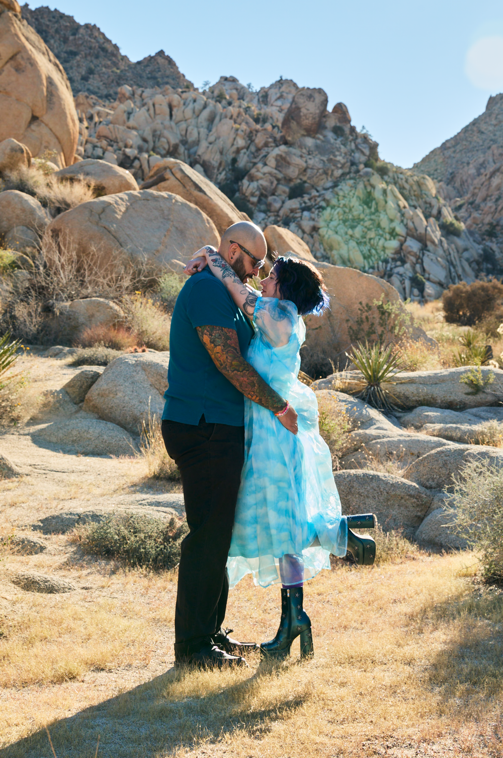 Couple sharing a kiss in Joshua Tree desert landscape, woman in blue dress, man with tattoos, rocky boulders behind.