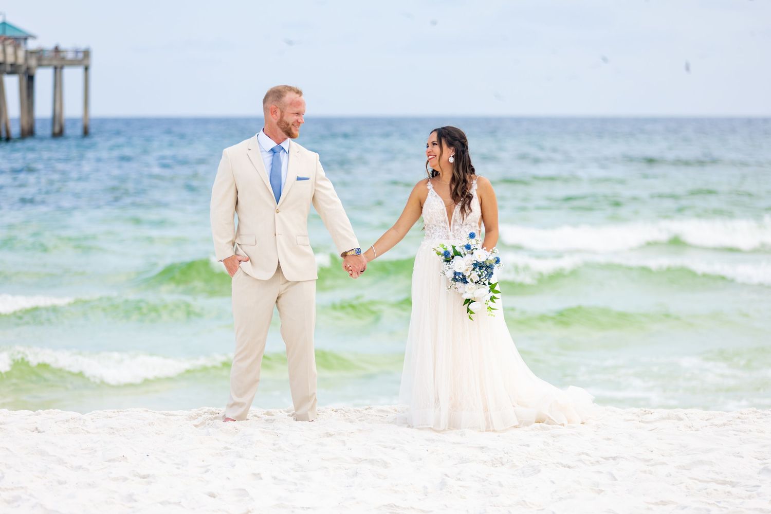 Bride and groom hold hands on a white sand beach, with turquoise ocean waves behind them on their wedding day.