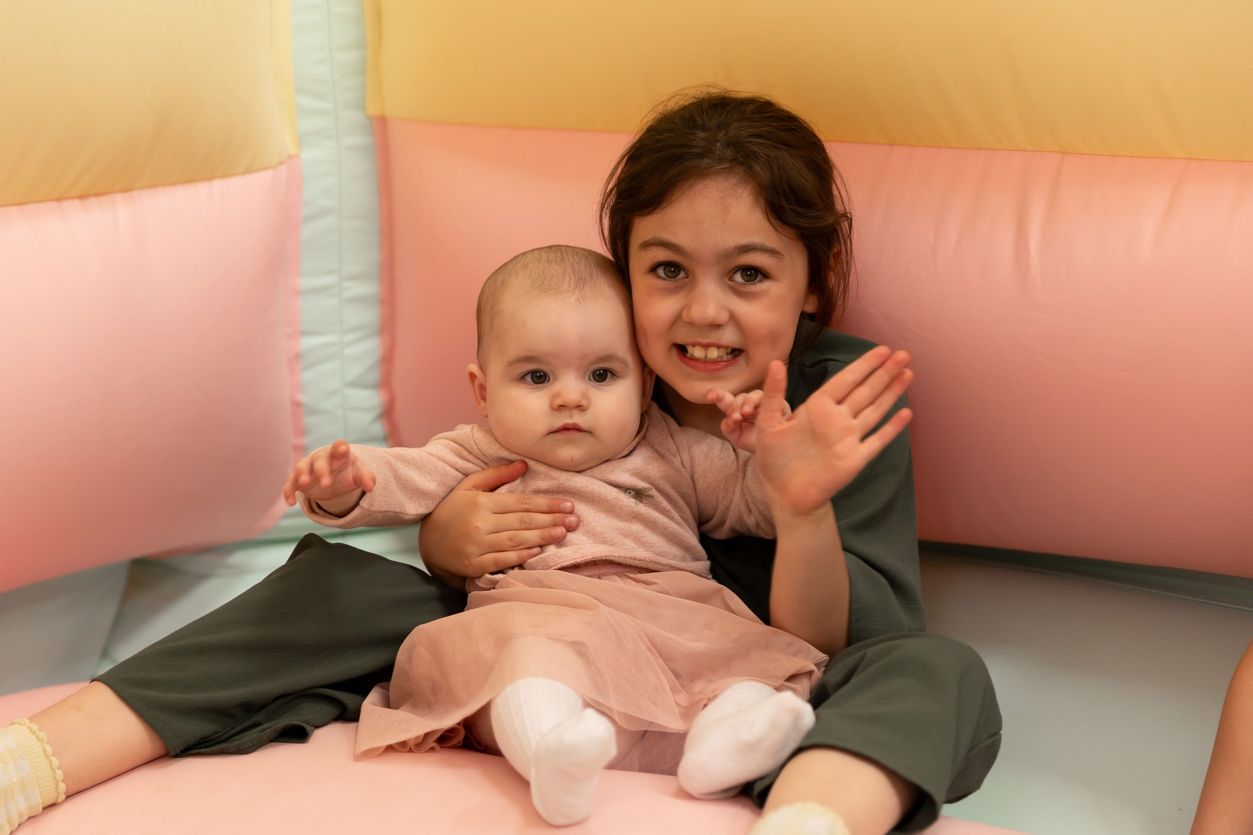 Older girl smiling and waving while holding a baby in a colorful soft play area.