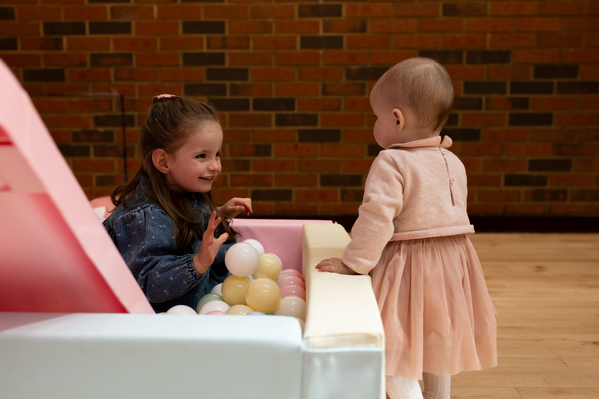 Two young girls playing together at a colorful ball pit toy in a bright indoor play area.