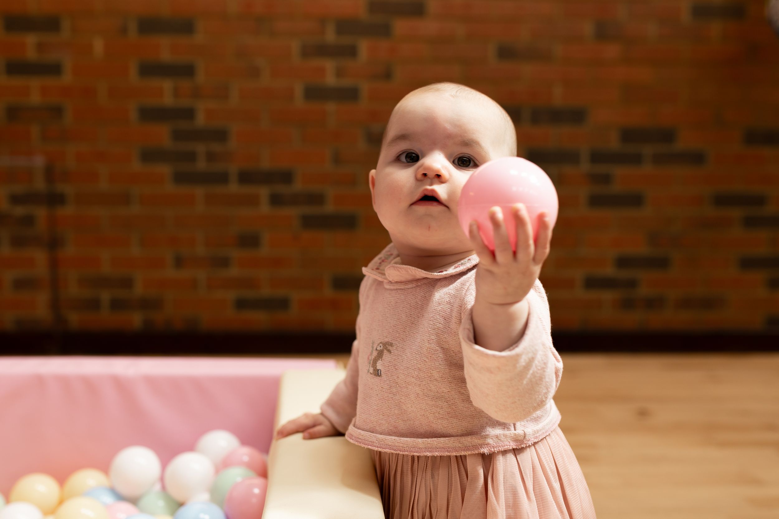 Baby girl in pink dress holding a pink ball in a colorful play area with brick wall background.