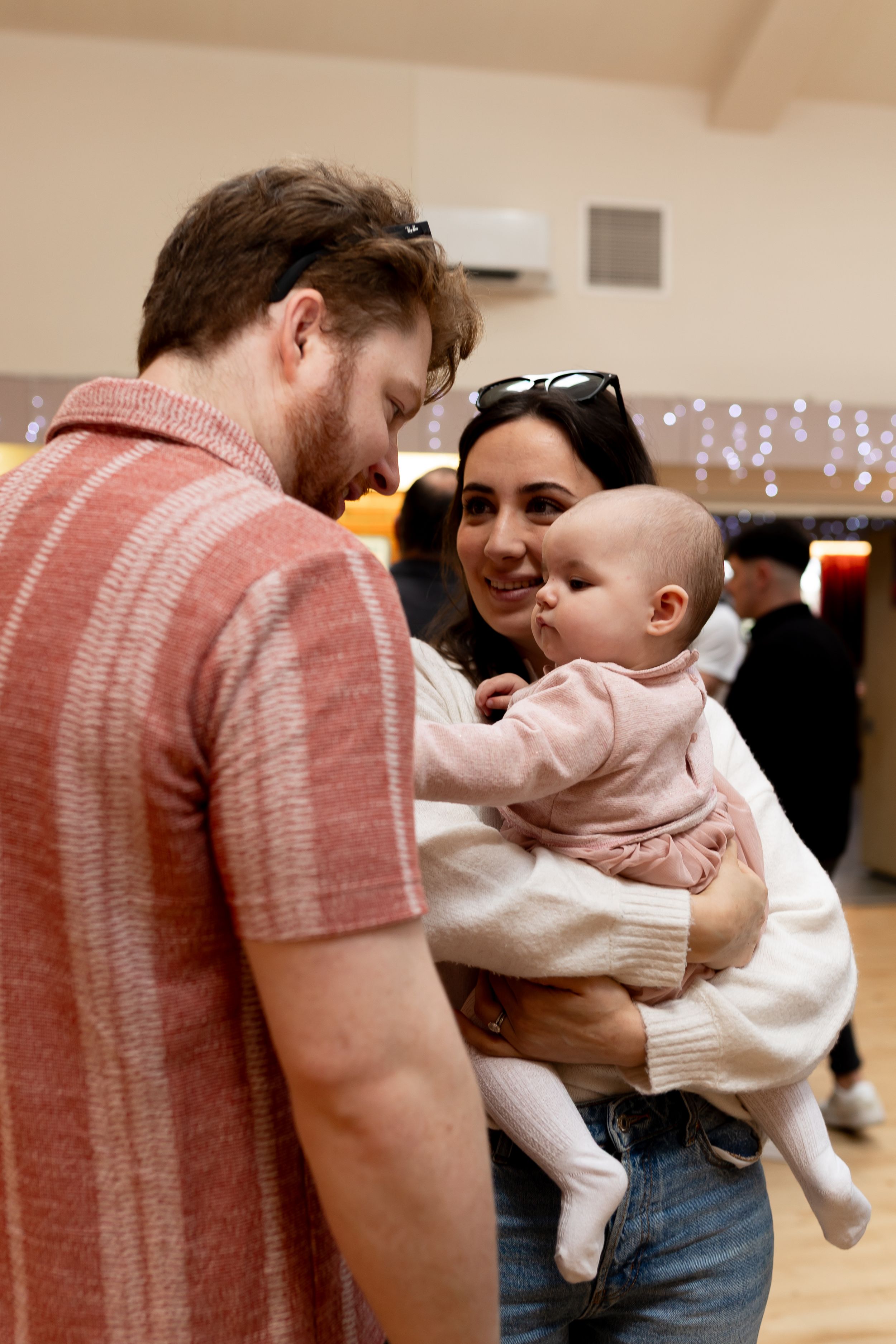A young couple smiling at their baby girl in a pink outfit at an indoor event.