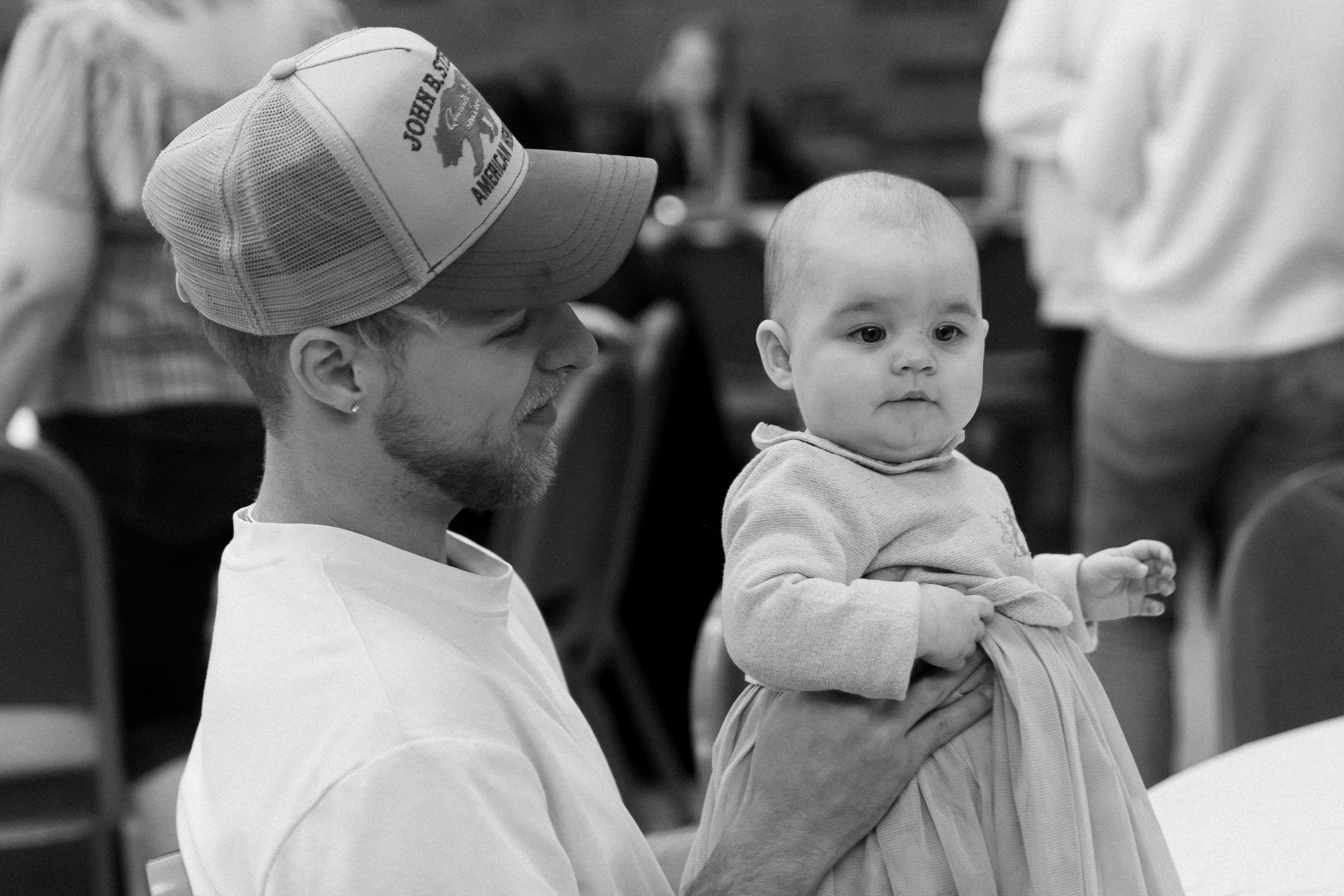 Black and white photo of a bearded man in a baseball cap tenderly holding a baby at an indoor gathering.