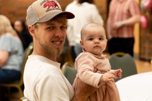 A smiling man wearing a trucker hat holds a cute baby in a pink outfit at an indoor family gathering.
