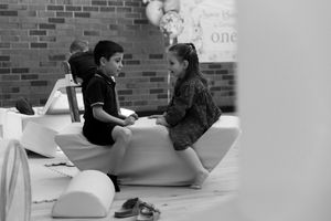 Two children sit facing each other on a white rocking toy inside a brick-walled room, captured in black and white.