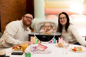 Happy family with baby in Joie high chair celebrating first birthday party at decorated table.