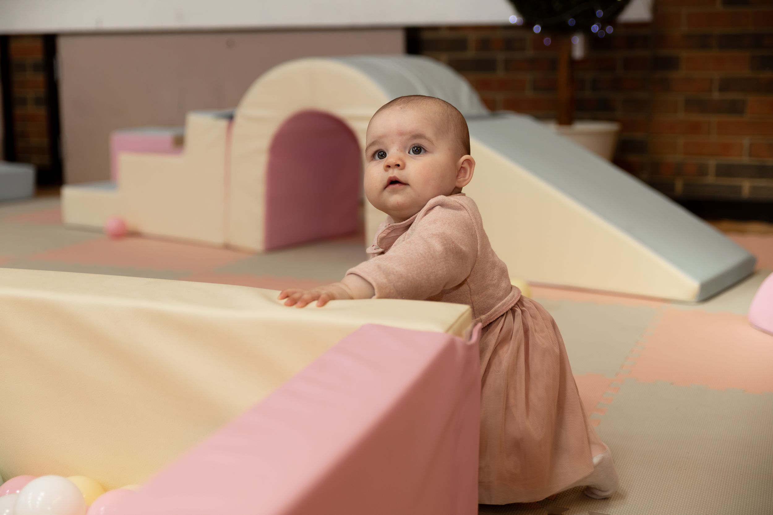 A curious baby girl in a pink dress plays in a soft play area with pastel foam shapes.