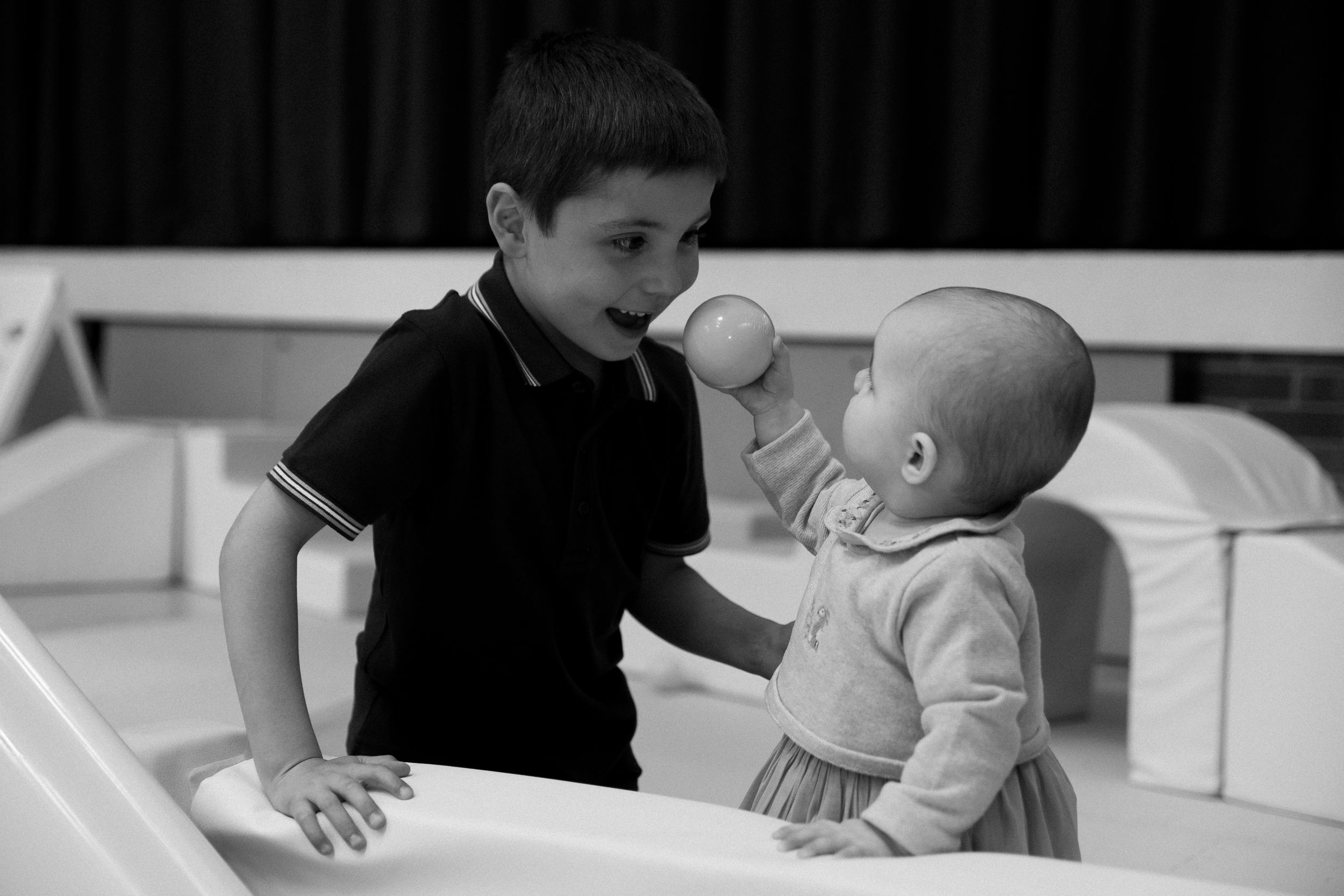 A young boy offers a ball to a baby in a soft play area in this black and white photo.