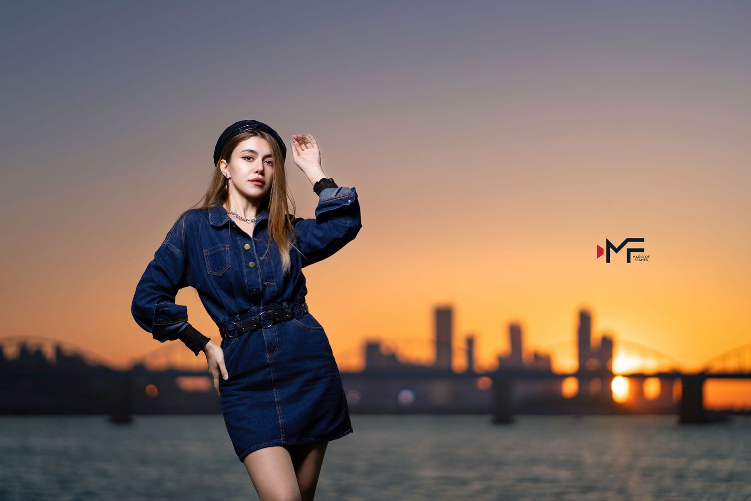 Young woman in denim dress poses at sunset with city skyline reflected over water in golden hour light.