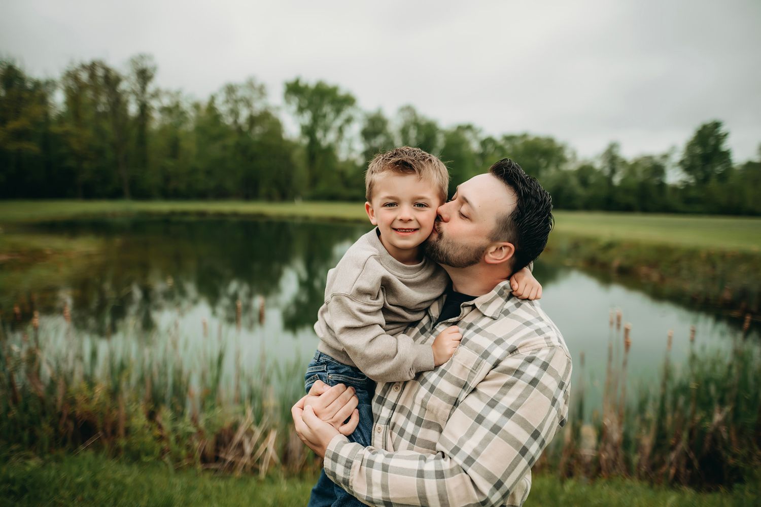 A father kisses his smiling young son on the cheek while holding him near a serene pond outdoors.