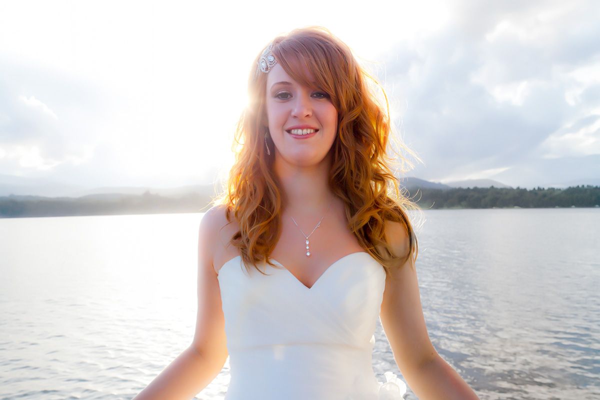 Smiling redhead bride in white strapless gown posing by a serene lake with soft sunlight and misty hills.