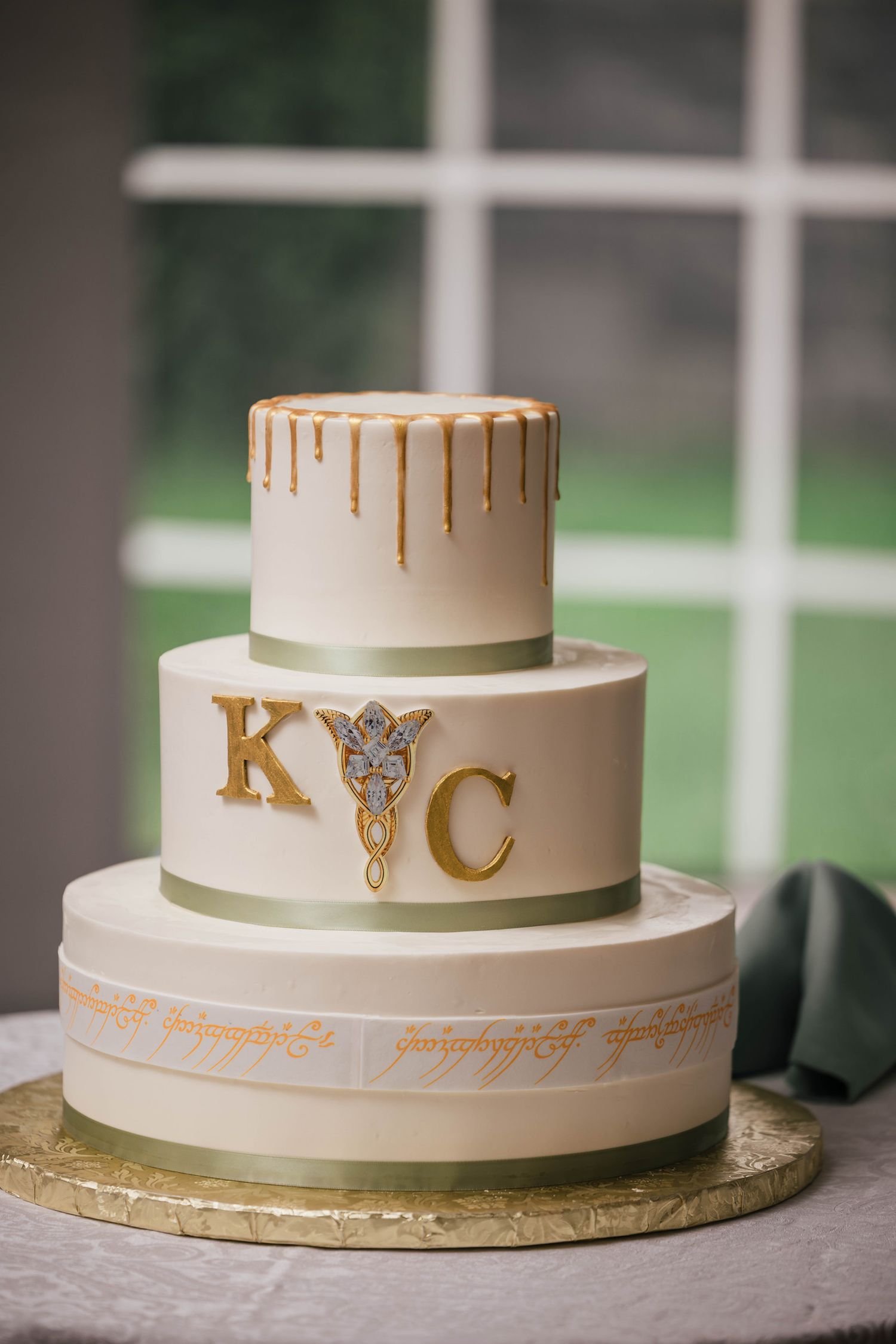 Three-tier wedding cake with gold drip icing, the couple's initials, an Evenstar pendant detail, and Elvish script, set by a window.