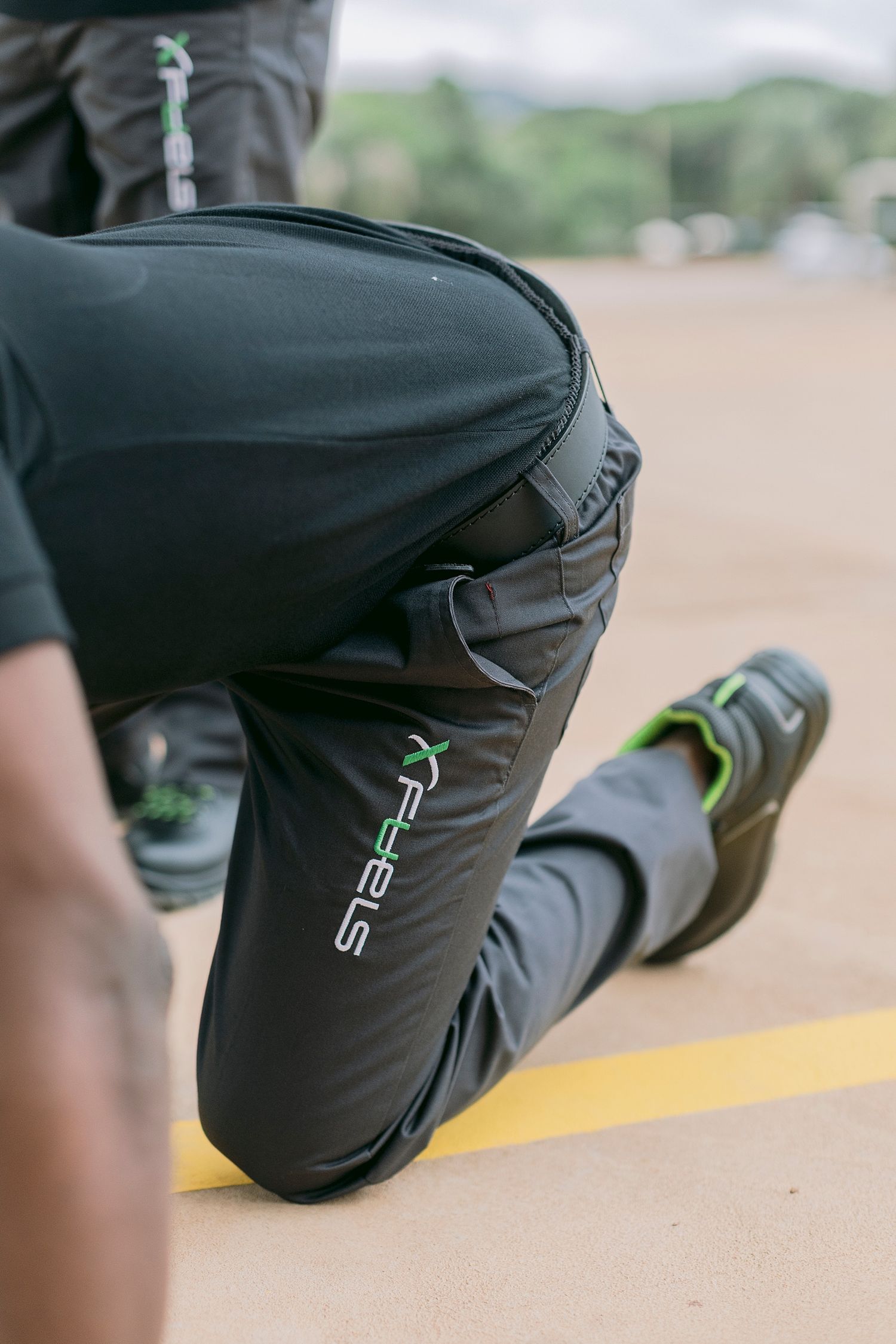 Athlete in black X-Feels athletic pants and green-accented shoes kneeling on a track surface.