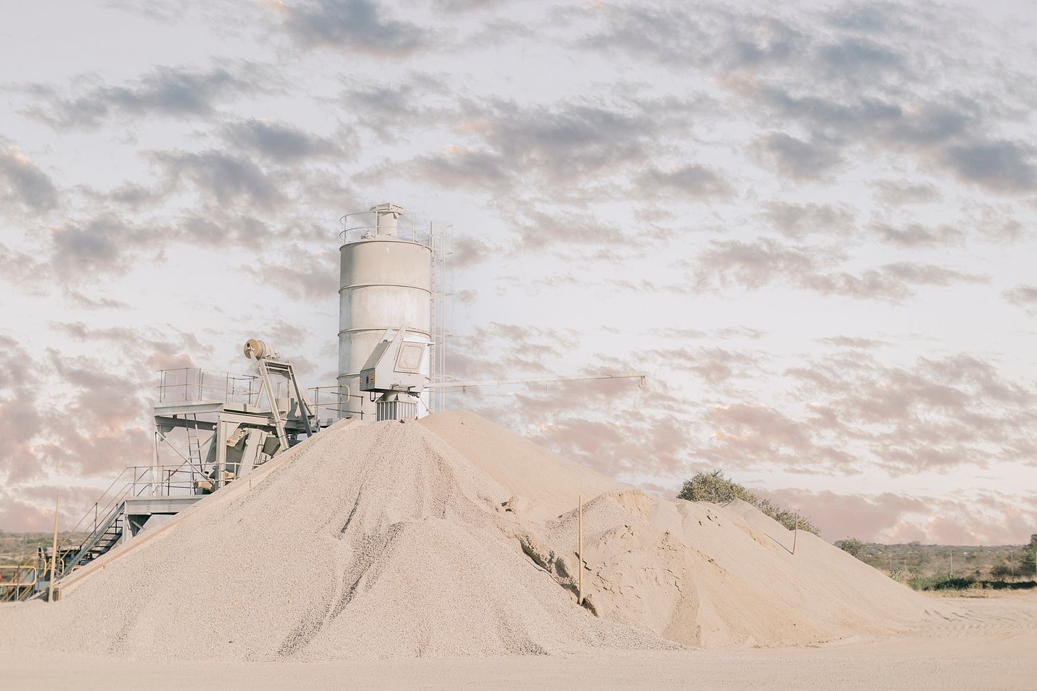 Large sand pile at a concrete batching plant with industrial silo and machinery under a cloudy sky.