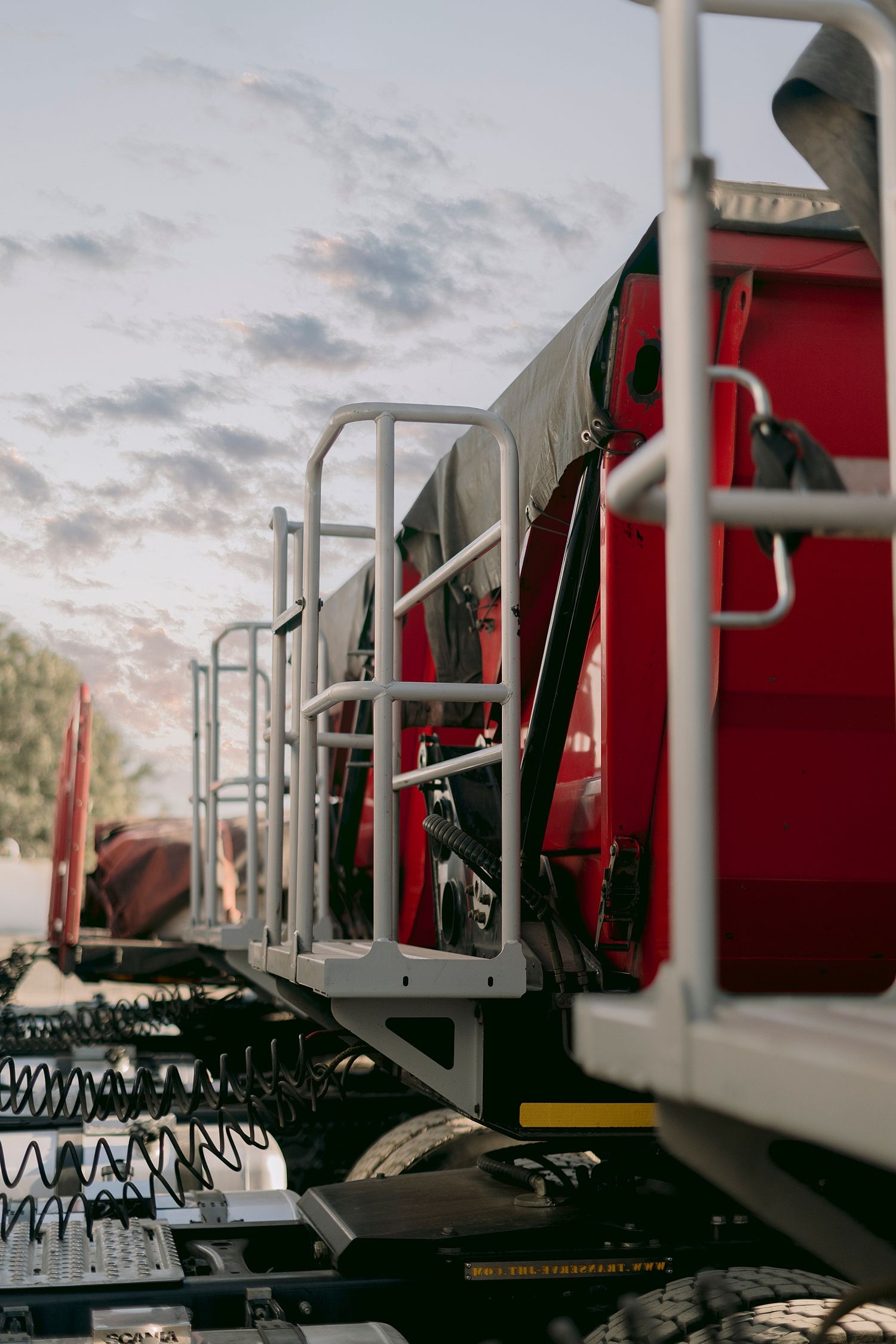 Close-up of a red semi-truck cab with white safety railings and tires visible against a cloudy sky.