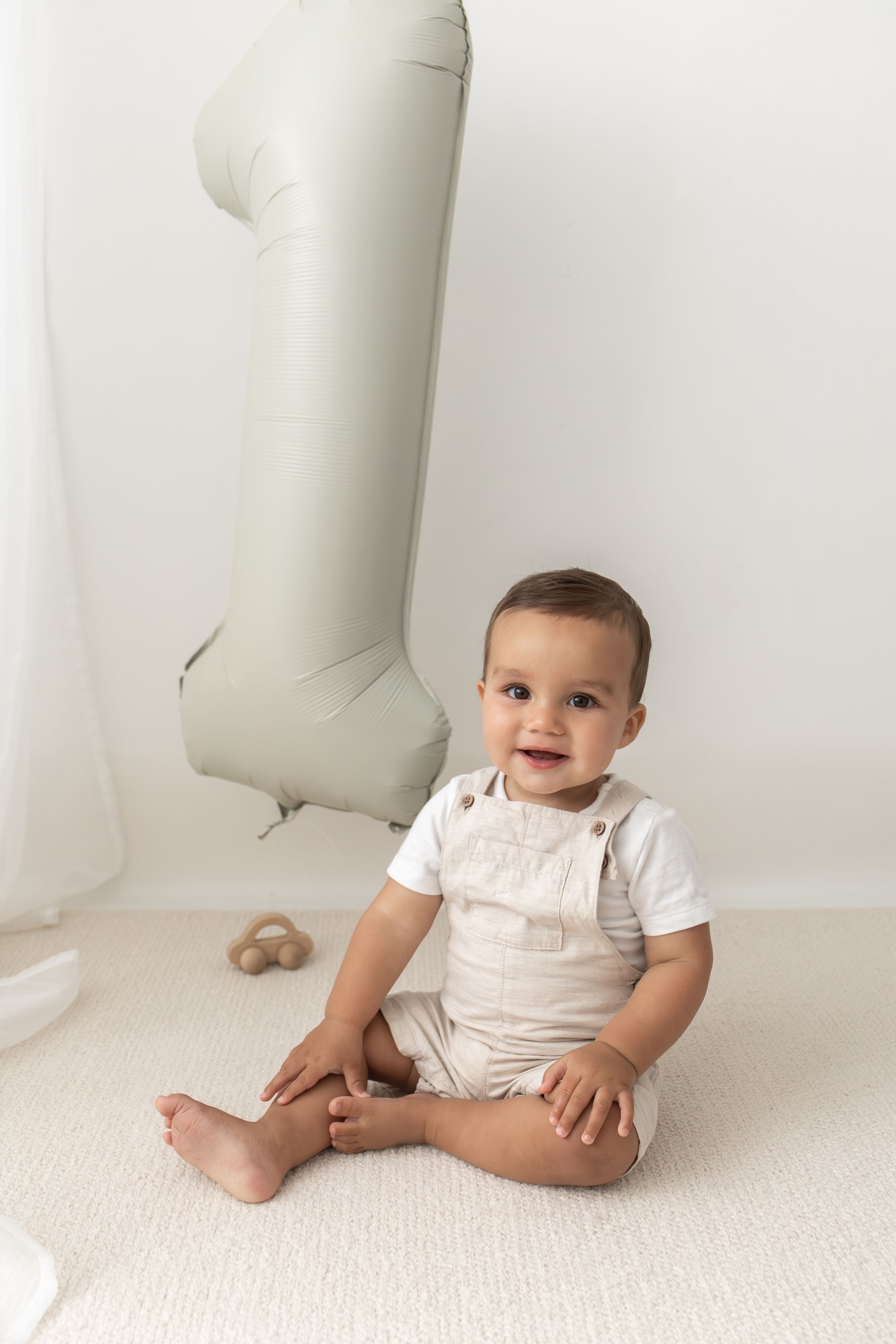 Smiling baby in beige sits on white carpet beside a large number 1 balloon for a first birthday natural birthday photoshoot Nottingham