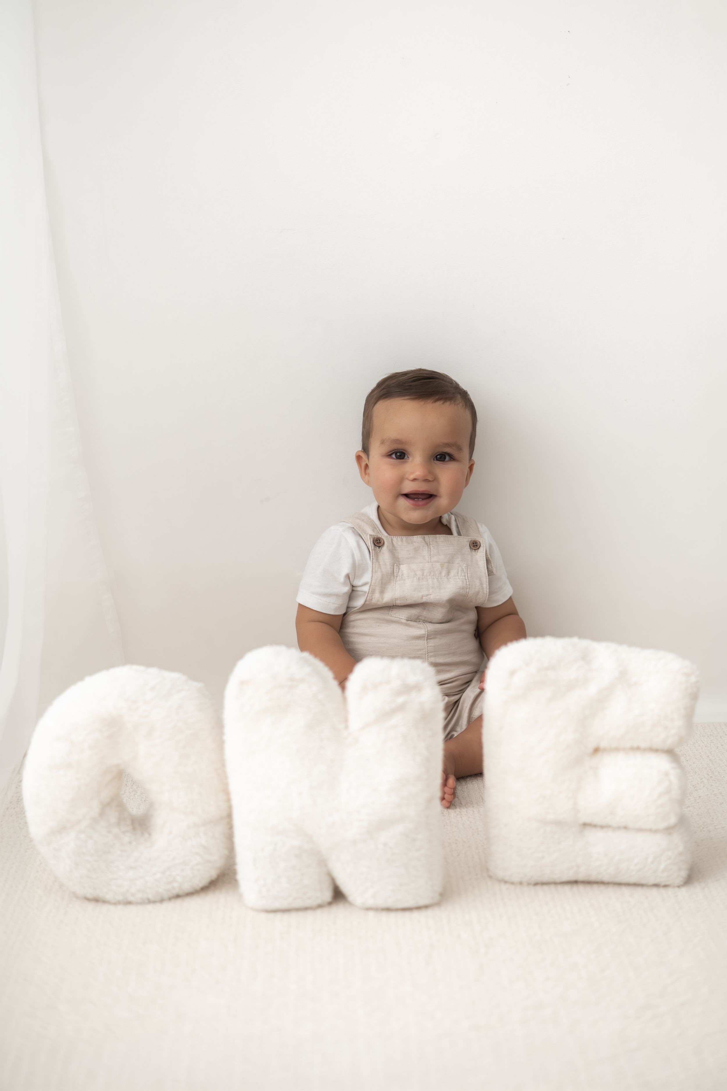 A smiling baby in a beige romper sits behind fluffy white letter pillows spelling ONE for a first birthday photo natural birthday photoshoot