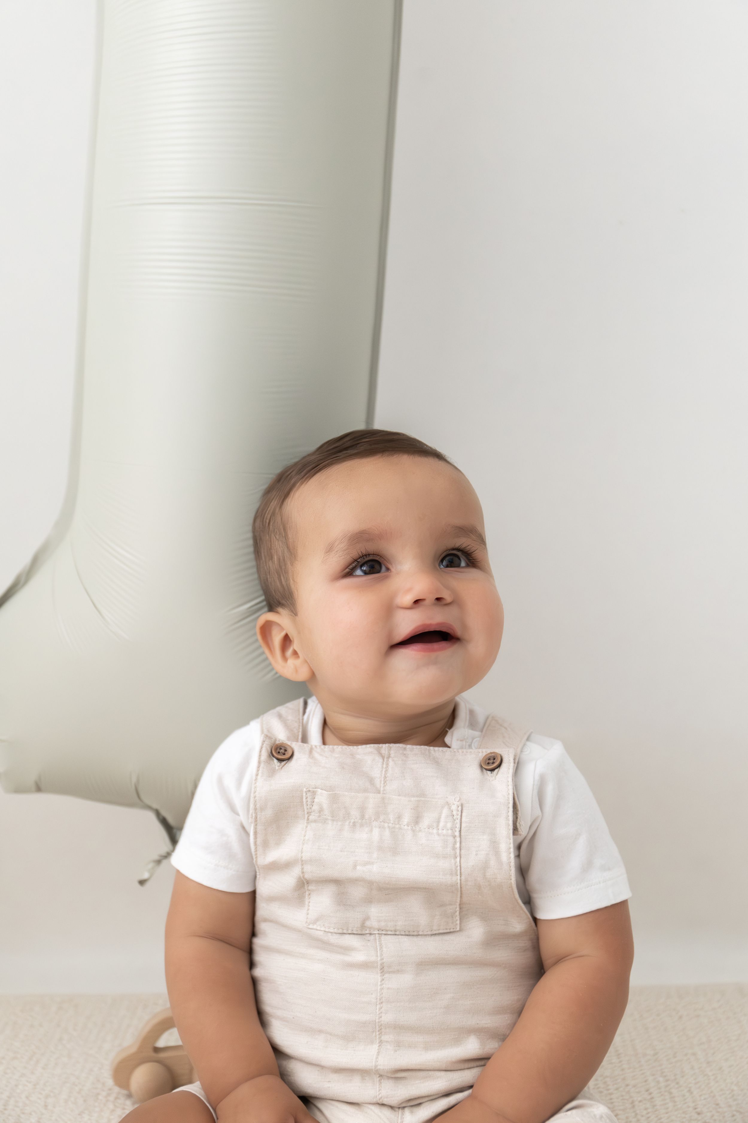Adorable baby in beige sitting on floor, smiling and looking up near a white 1 balloon natural 1st birthday photoshoot Nottingham