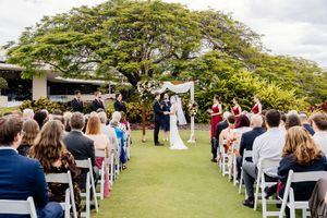 Outdoor wedding ceremony on green lawn with guests seated in white chairs beneath a large tropical tree in Brisbane Queensland