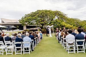 Outdoor wedding ceremony on lush green lawn with white chairs, floral arch, and large tree backdrop under cloudy sky  in Brisbane Queensland