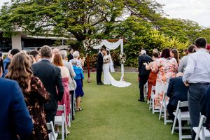 Outdoor wedding ceremony on green lawn with guests seated in white chairs facing couple at floral arch at Indooroopilly Golf Club Brisbane