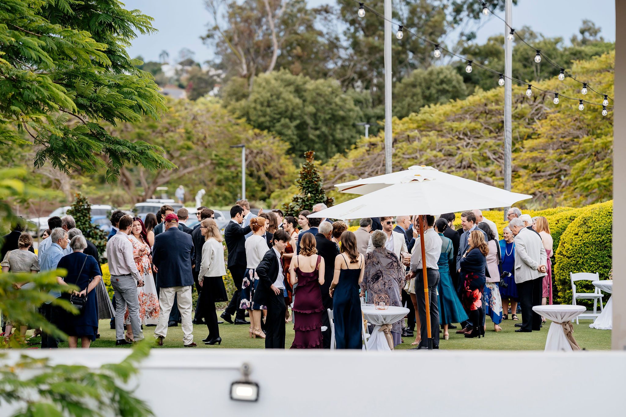 Elegant outdoor wedding reception gathering on lush green lawn with guests mingling under white umbrellas surrounded by trees in Queensland